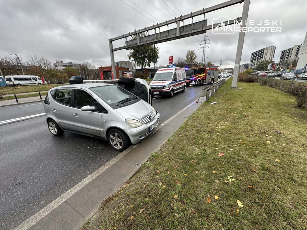 Na drodze stoi przewrócony samochód osobowy, obok widać karetki i straż pożarną, w tle ruch uliczny.
