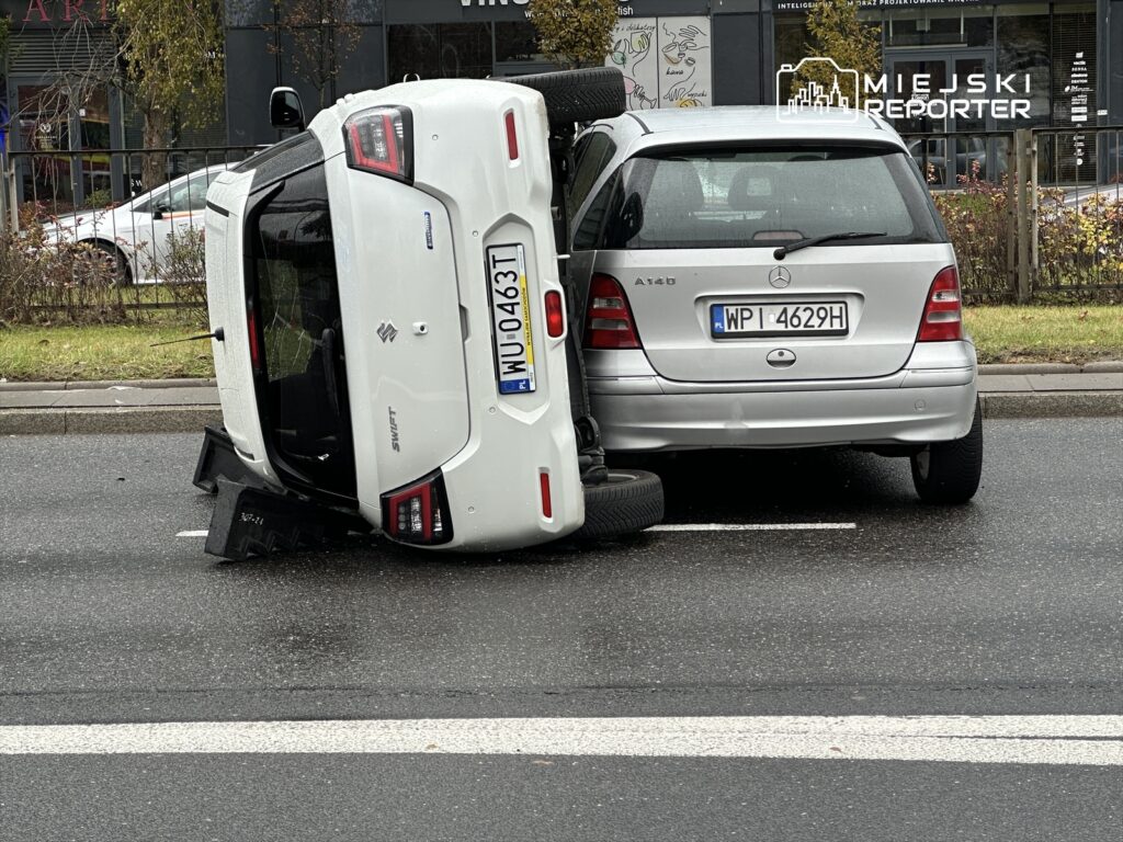 Biały samochód osobowy leży na boku obok srebrnego pojazdu na miejskiej ulicy.