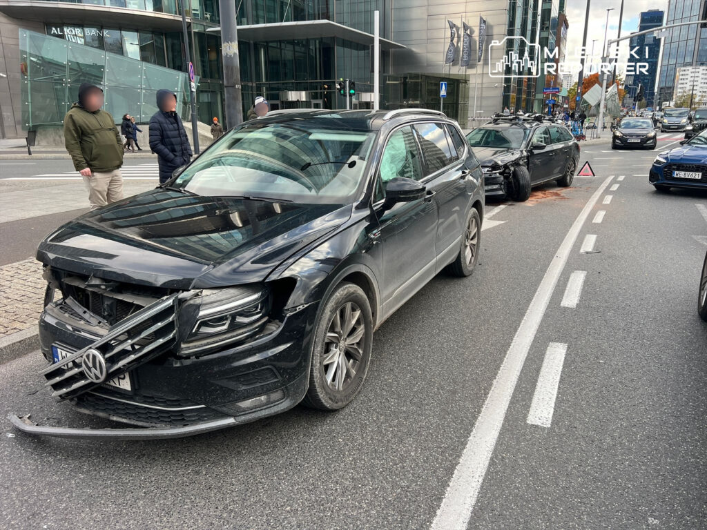 Dwa uszkodzone samochody osobowe na ruchliwej ulicy, obok stojący mężczyzna w kurtce i dwóch innych obserwatorów.
