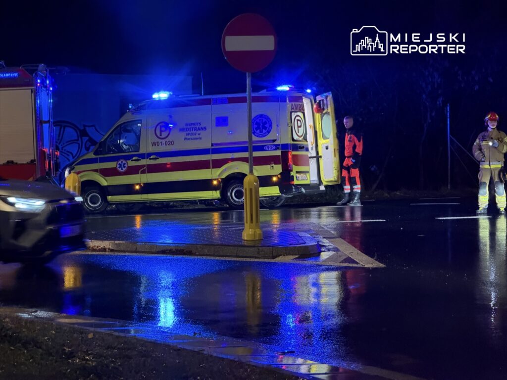 Karetka pogotowia z włączonymi sygnałami świetlnymi stoi na drodze, obok niej ratownicy w odzieży ochronnej.