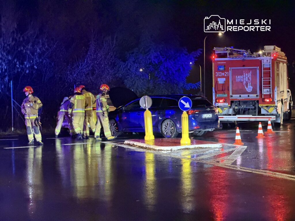 Grupa strażaków w hełmach bada uszkodzony samochód osobowy na mokrej drodze, obok stoi wóz strażacki.