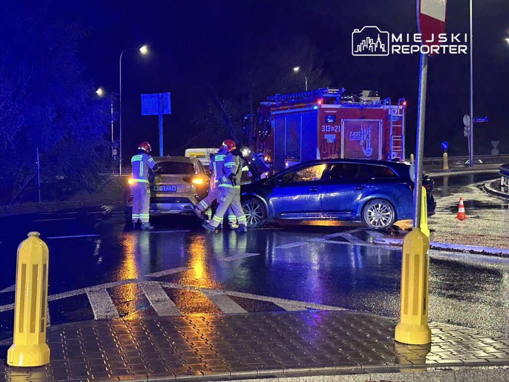 Trzech strażaków w kaskach bada uszkodzone samochody na mokrej jezdni w nocy, obok wozu strażackiego.