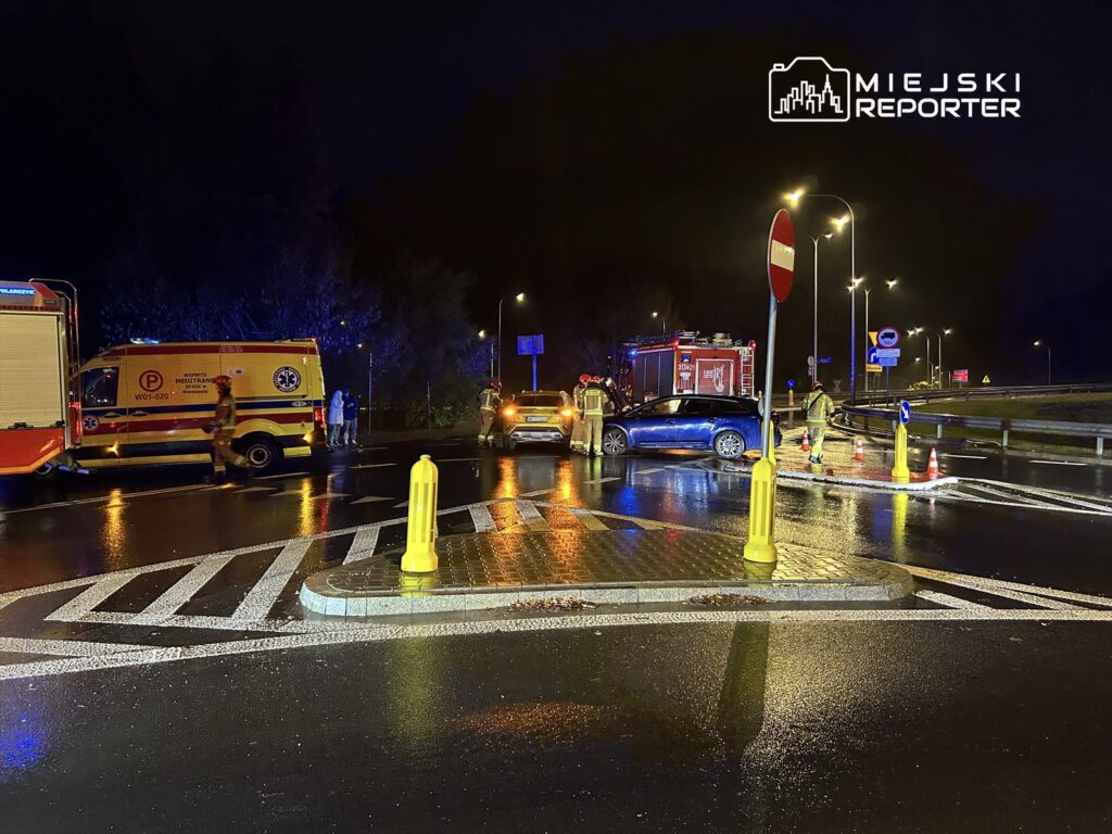 Na skrzyżowaniu w nocy, zespół ratunkowy udziela pomocy po wypadku drogowym; w tle widoczne pojazdy służb.