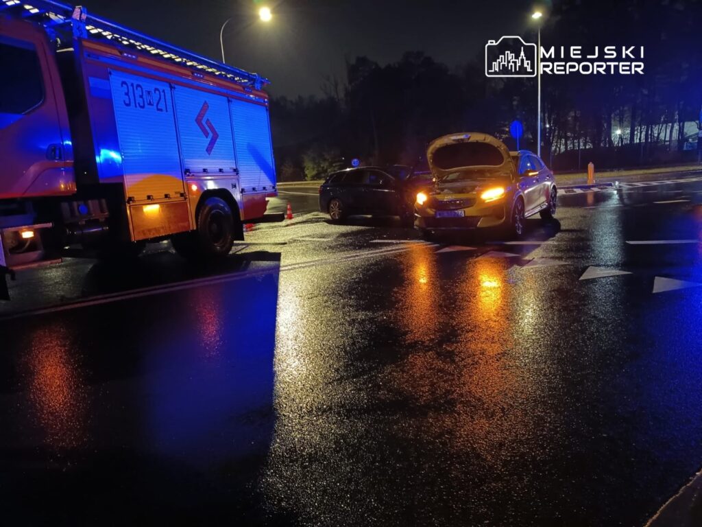 W nocy na mokrej jezdni stoi żółty samochód z otwartą maską, obok niego znajduje się wóz strażacki z włączonymi sygnałami.