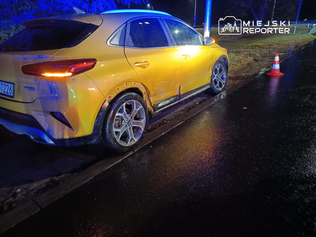 Żółty samochód osobowy zaparkowany na mokrej nawierzchni, obok trójkąt ostrzegawczy w nocy.