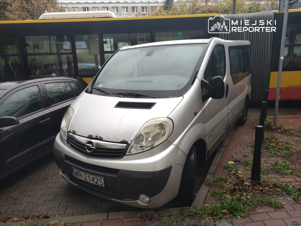Srebrny van zaparkowany na wąskiej ulicy, obok czarnego samochodu, w tle żółty autobus.