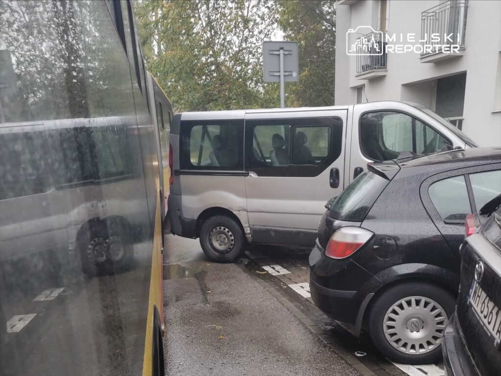 Biały bus zaparkowany w wąskiej uliczce, z trzech stron otoczony osobowymi samochodami.