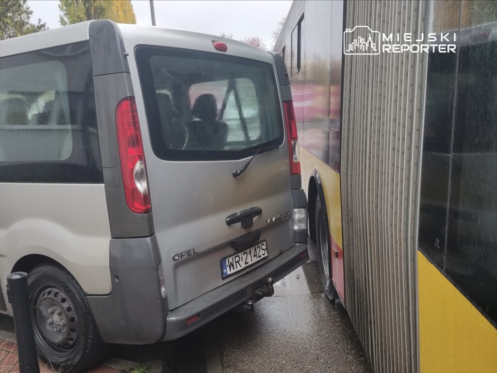 Szary samochód dostawczy zaparkowany blisko autobusu miejskiego, obydwa pojazdy stoją na mokrej nawierzchni.