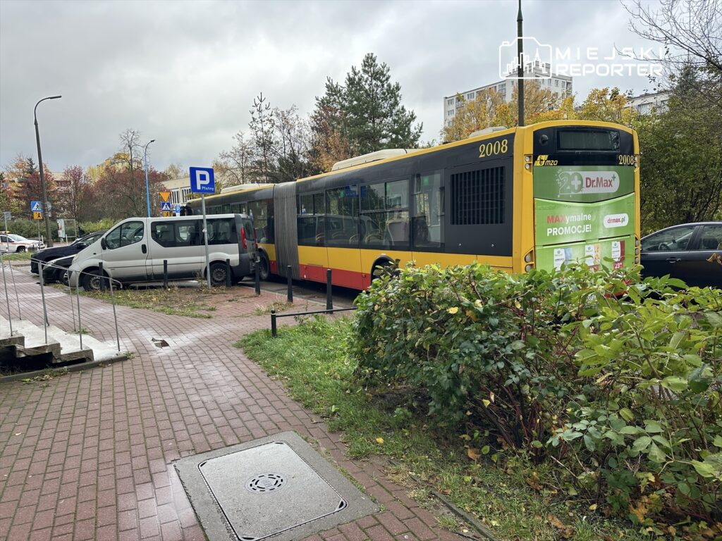 Autobus miejski zatrzymany obok zaparkowanych samochodów na chodniku, w otoczeniu drzew i krzewów.