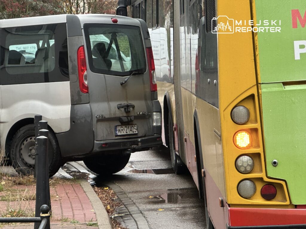 Samochód osobowy zaparkowany blisko autobusu na wąskiej ulicy, obok mokrej nawierzchni i metalowych słupków.