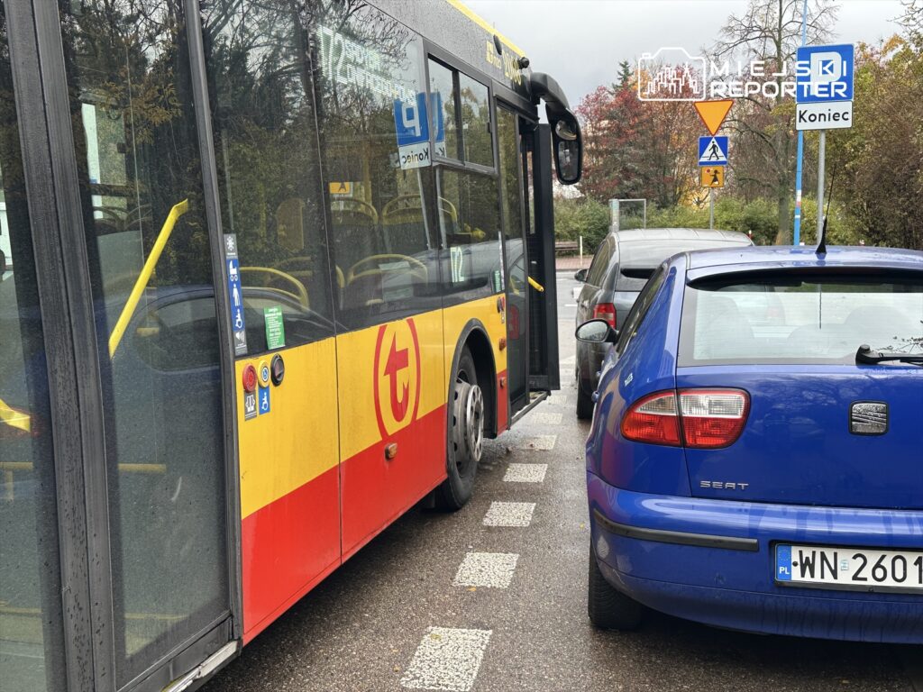 Autobus komunikacji miejskiej zatrzymany obok zaparkowanego nieprawidłowo niebieskiego samochodu osobowego.