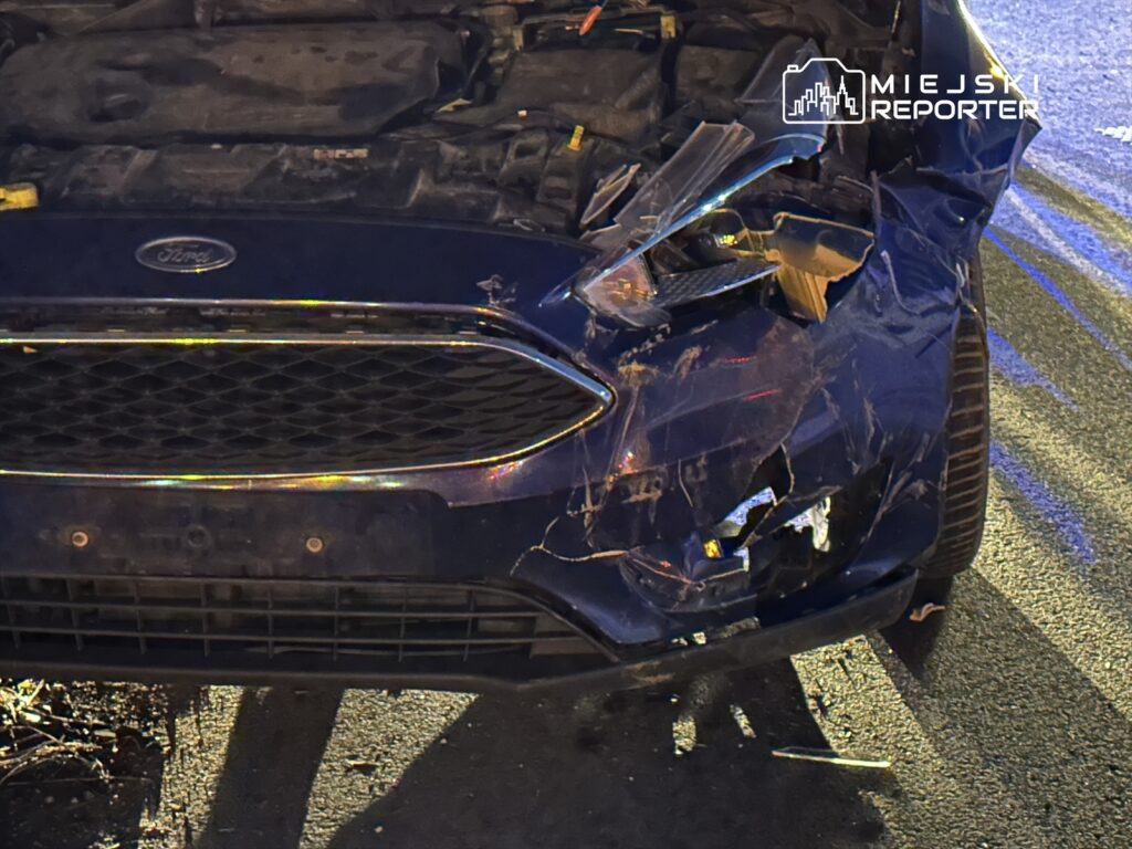 Uszkodzony niebieski samochód osobowy z wgniecionym przodem stoi na drodze, obok widoczna nawierzchnia asfaltowa.