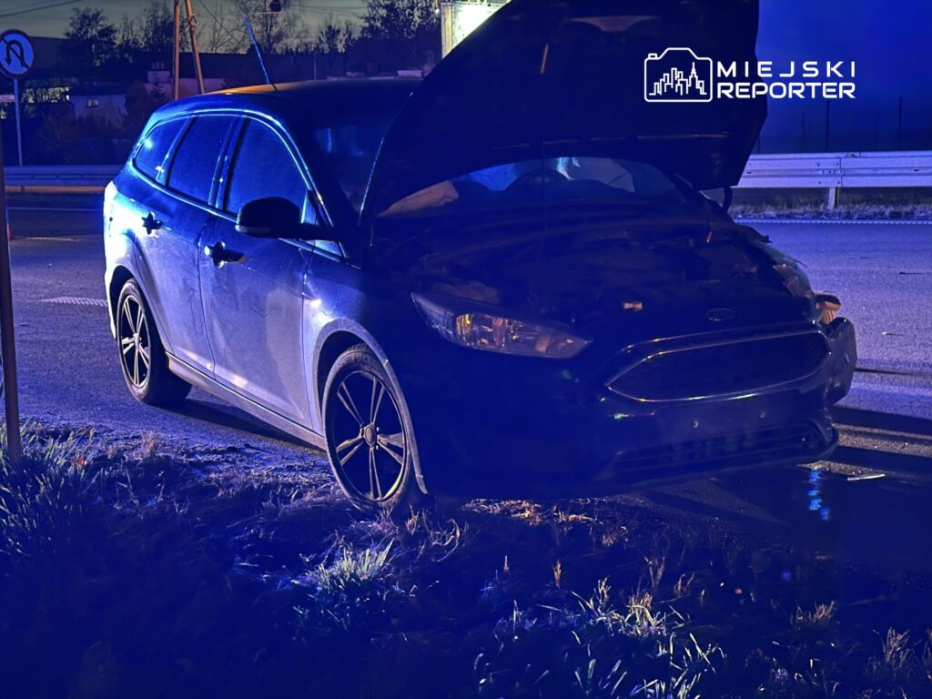 Czarny samochód osobowy z otwartą maską stoi na poboczu drogi w świetle niebieskich reflektorów.