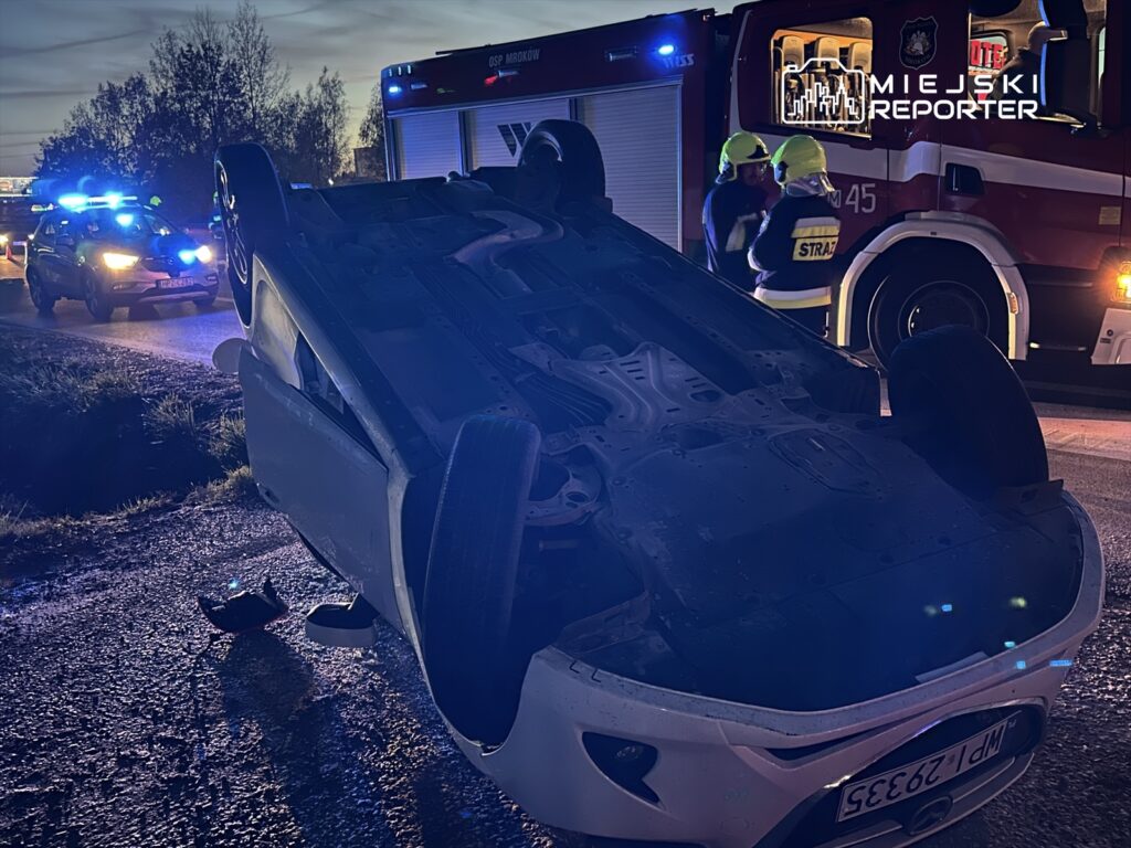Przewrócony samochód osobowy leży na drodze, a w tle strażacy rozmawiają obok wozu strażackiego z włączonymi sygnałami.