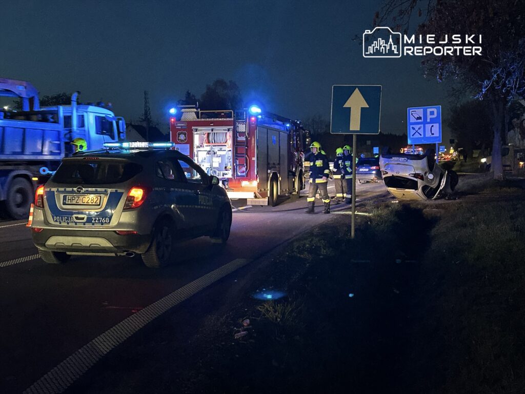 Na drodze w nocy, radiowóz i wóz strażacki, obok przewrócone auto, a strażacy oceniają sytuację.