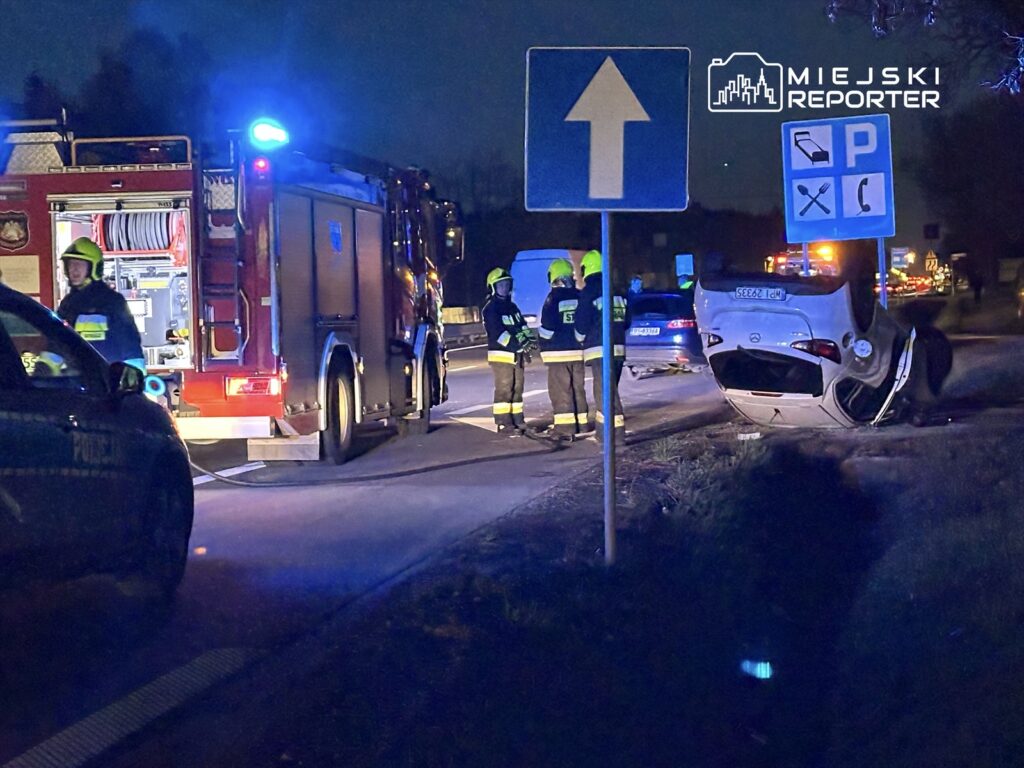 Strażacy w kaskach pomagają przy przewróconym samochodzie na drodze, w tle widać policyjny radiowóz i inne pojazdy.