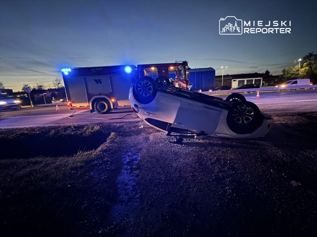 Na drodze stoi przewrócone na dach białe auto, obok widać woz strażacki z włączonymi sygnałami świetlnymi.