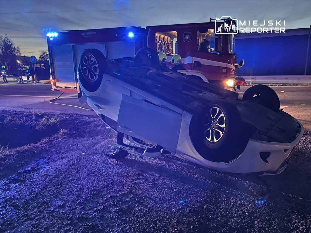 Odwrócony samochód osobowy leży na poboczu drogi, obok działa straż pożarna z włączonymi sygnałami świetlnymi.