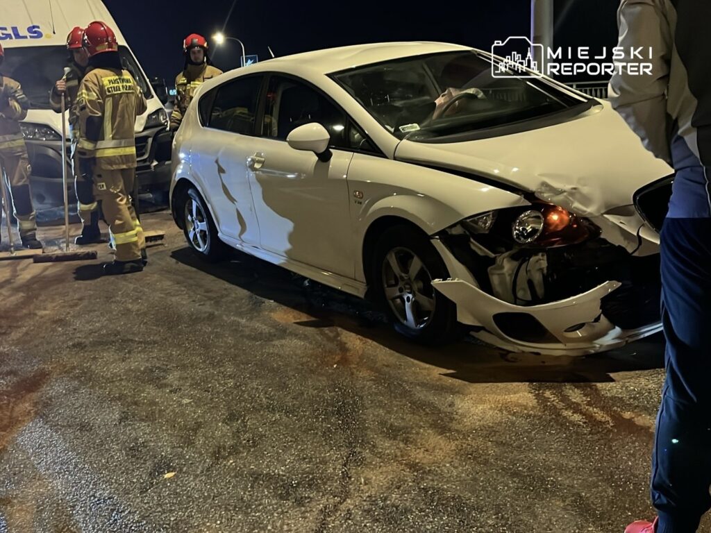 Czterech strażaków w mundurach omawia sytuację obok uszkodzonego białego samochodu osobowego na drodze nocą.