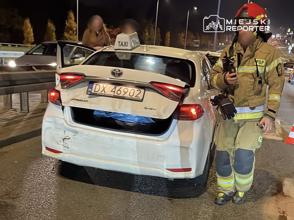 Biały samochód taxi z uszkodzeniami stoi na drodze, obok strażak w mundurze z radiotelefonem.