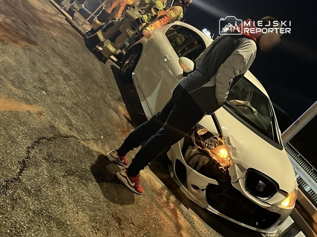 Mężczyzna w dresie stoi obok uszkodzonego białego samochodu na drodze, w tle strażacy w mundurach.