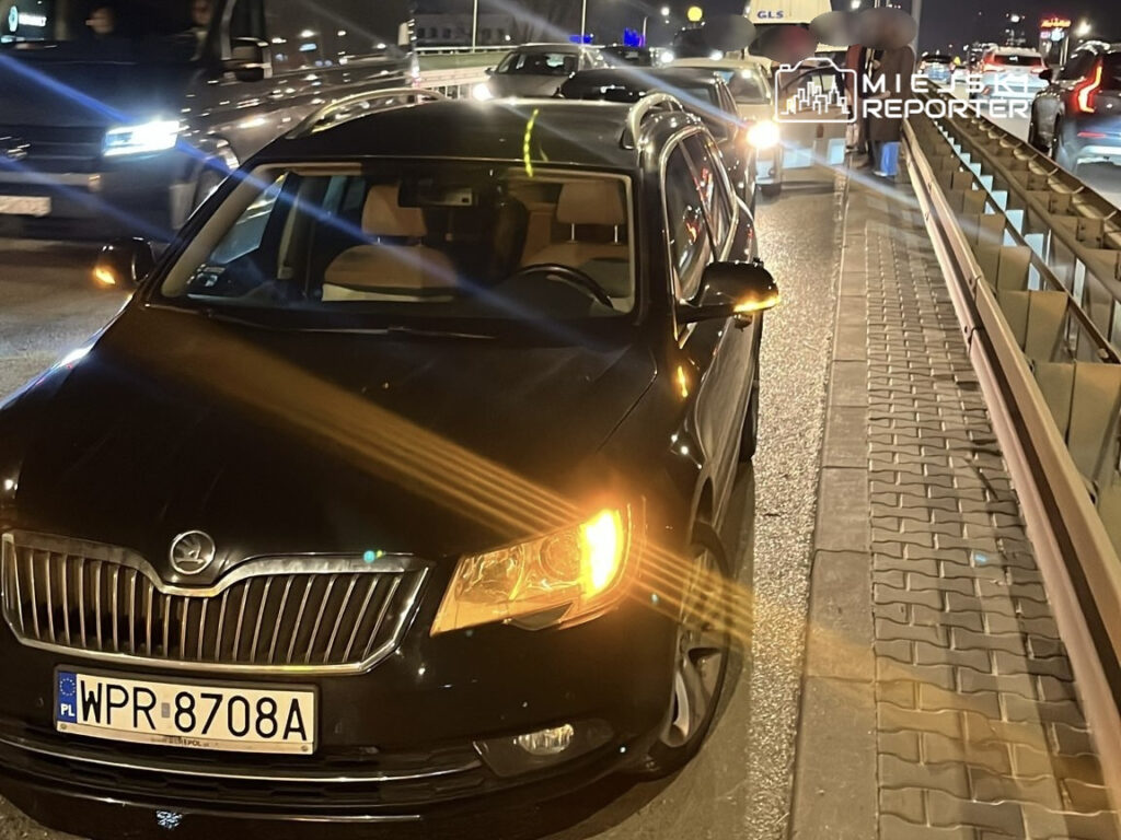 Czarny samochód osobowy zatrzymany na pasie ruchu w nocy, obok innych pojazdów na ruchliwej drodze.