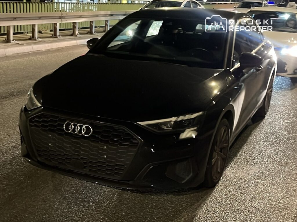 Czarny samochód marki Audi zaparkowany na oświetlonej ulicy w nocy, w otoczeniu innych pojazdów.