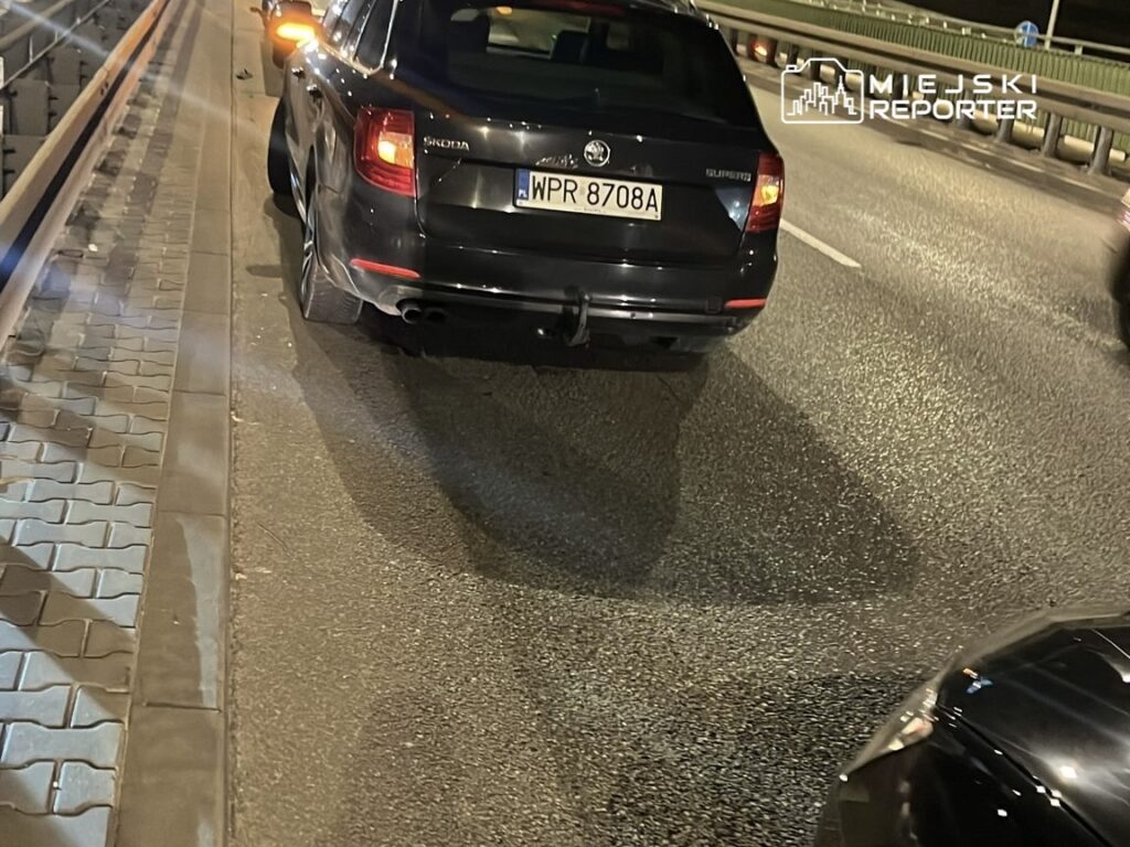 Czarny samochód osobowy stojący na pasie ruchu, obok widoczne inne pojazdy na oświetlonej ulicy.