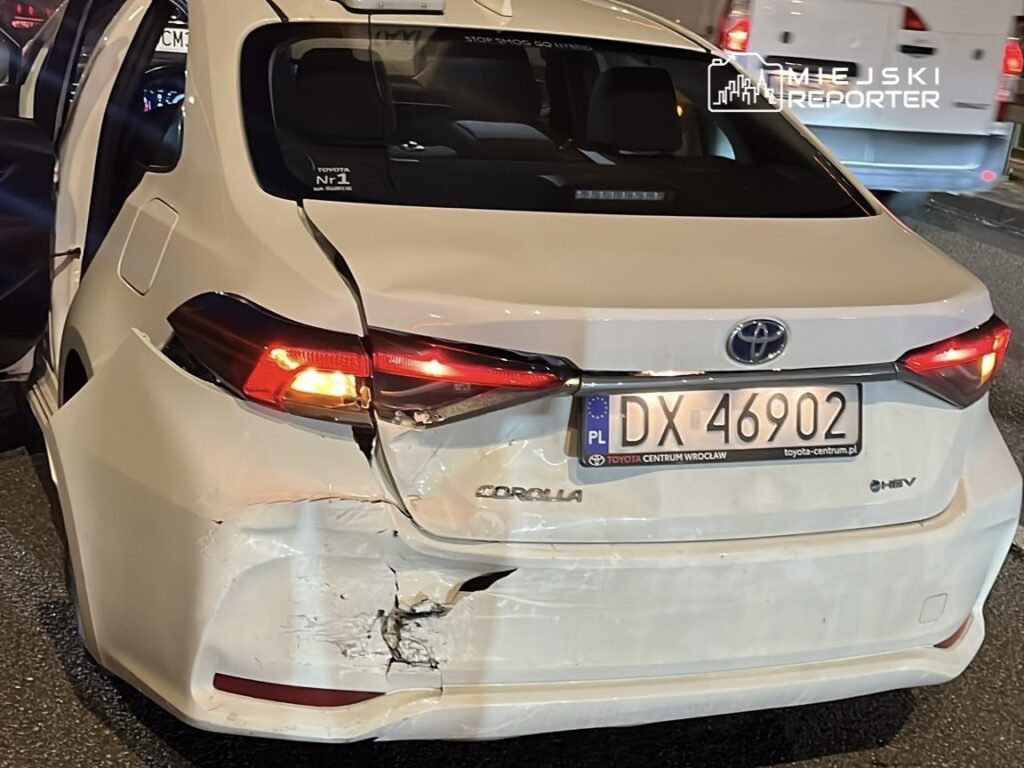 Uszkodzony biały samochód osobowy marki Toyota Corolla z widocznymi wgnieceniami na tylnej części, zaparkowany na ruchliwej ulicy.