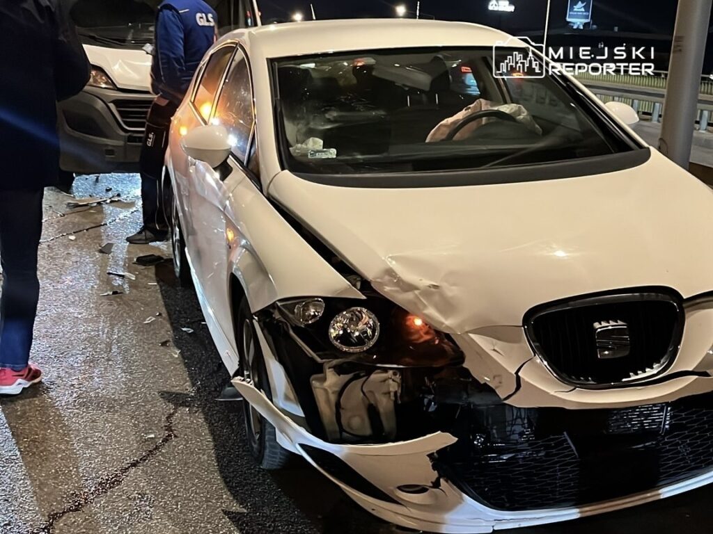 Biały samochód osobowy z uszkodzoną przednią częścią stoi na jezdni, obok dwóch mężczyzn rozmawiających w nocy.