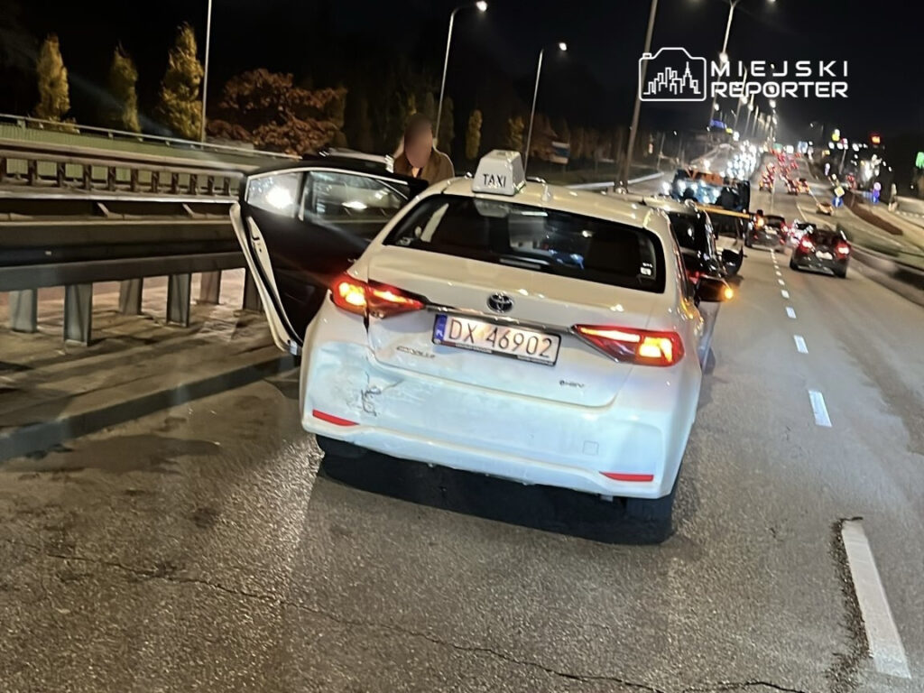 Taksi zatrzymane na jezdni nocą, kierowca wysiada z pojazdu, a w tle widać ruch uliczny.