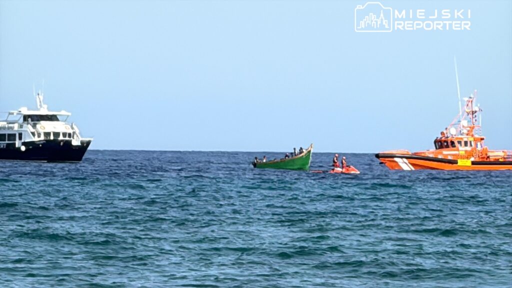 Zielona łódź z kilkoma osobami na pokładzie jest otoczona przez ratunkowy statek i motorówkę na spokojnym morzu.