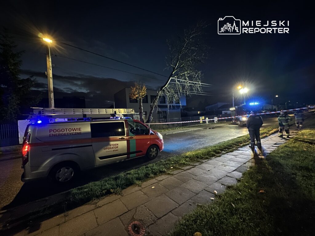 Samochód pogotowia energetycznego z włączonymi sygnałami świetlnymi na ulicy, obok dwóch pracowników w odblaskowych kamizelkach.