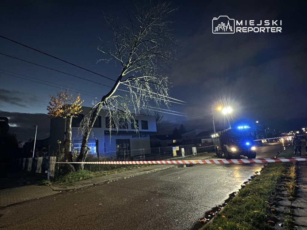 Na ulicy w nocy stoi wóz strażacki z włączonymi światłami, a teren jest ogrodzony taśmą zabezpieczającą.