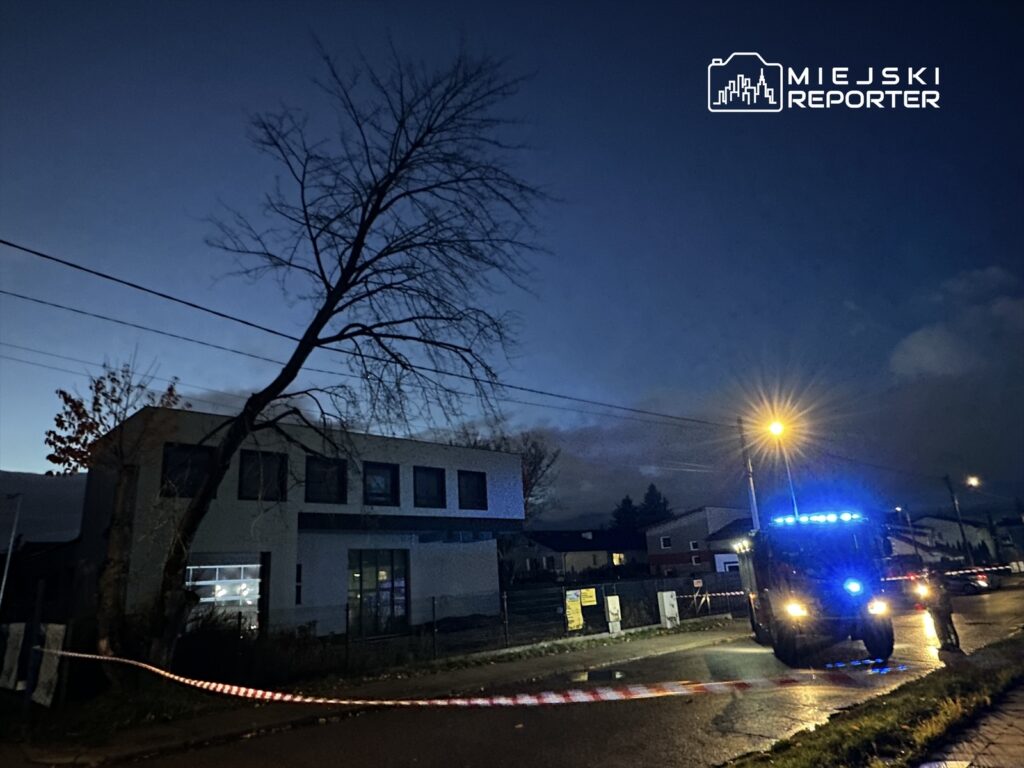 Policyjny radiowóz z niebieskimi światłami stoi obok nowoczesnego budynku w zmierzchowym otoczeniu, zabezpieczając teren.