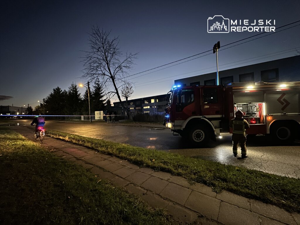 Strażak obok wozu strażackiego na oświetlonej ulicy wieczorem, podczas akcji ratunkowej w okolicy budynków.