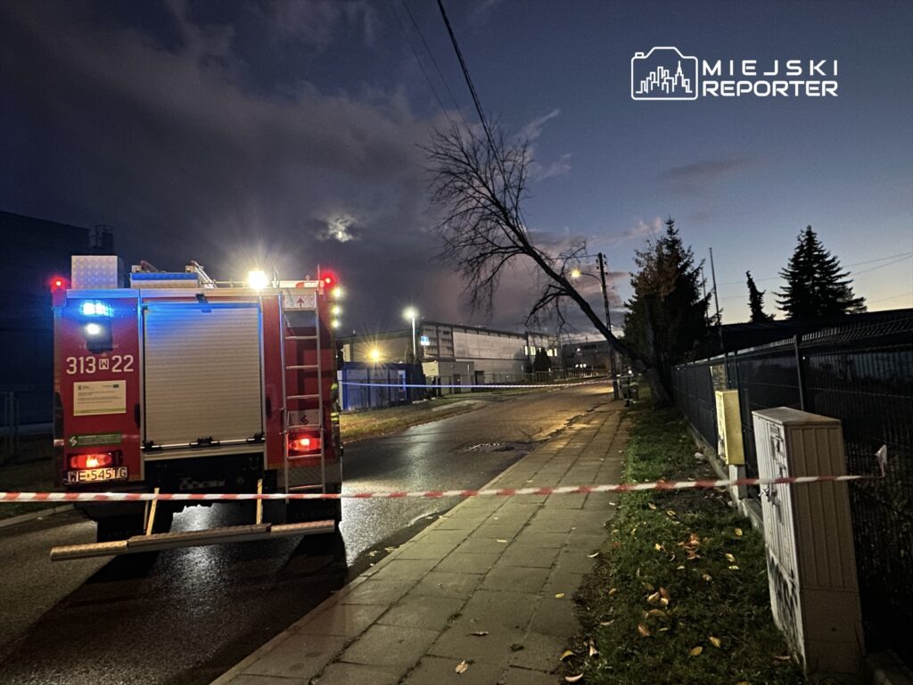 W tle widać woz strażacki z włączonymi sygnalizatorami, zaparkowany na oświetlonej ulicy.