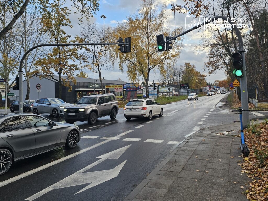 Ruch uliczny na skrzyżowaniu z zielonym światłem, w tle budynki i reklamy, jesienne liście na jezdni.