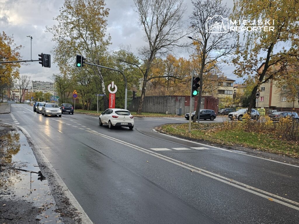 Ruch samochodowy na skrzyżowaniu z zielonym światłem, otoczone drzewami w jesiennych kolorach.