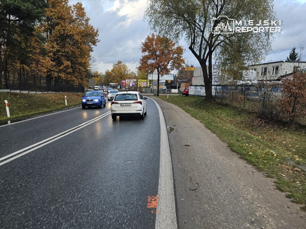 Ruch samochodowy na drodze z widocznymi drzewami w jesiennych kolorach oraz billboardami w tle.