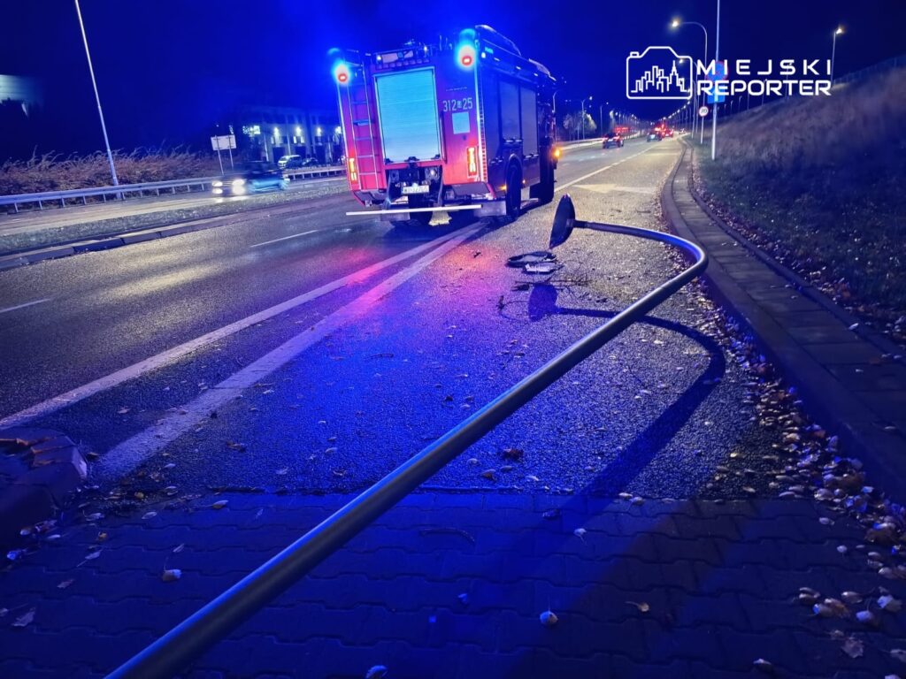 W tle widać woz strażacki z włączonymi sygnalizatorami na oświetlonej ulicy, obok leży złamany słup.