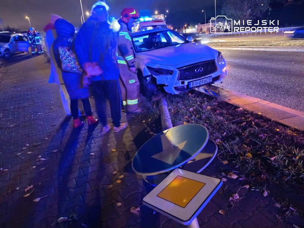 Grupa osób obserwuje działania ratowników przy uszkodzonym samochodzie na poboczu drogi, otoczonym przez niebieskie światła.