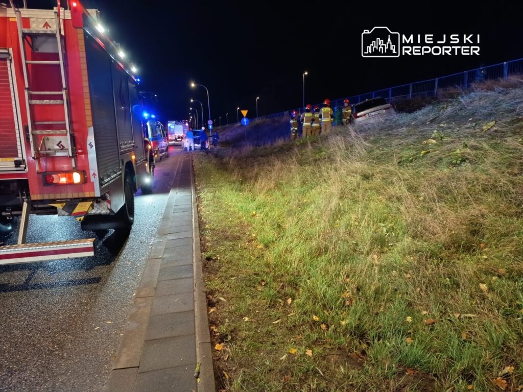 W nocy na poboczu drogi, strażacy i ratownicy pracują przy wypadku samochodowym, obok zjeżdżającej karetki.