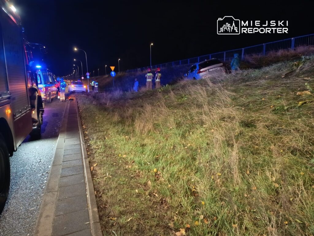 Na poboczu drogi stoją strażacy i policjanci, obok przewrócone auto oraz pojazdy służb ratunkowych.