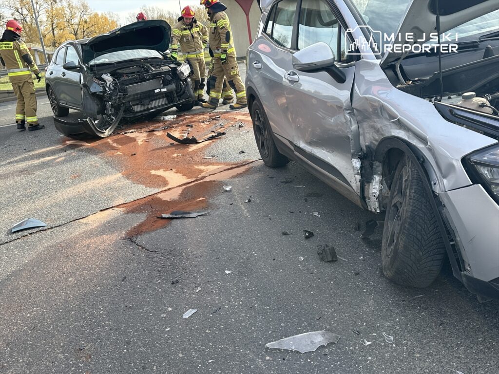 Cztery osoby w mundurach strażackich pracują przy uszkodzonych samochodach po wypadku drogowym na jezdni.