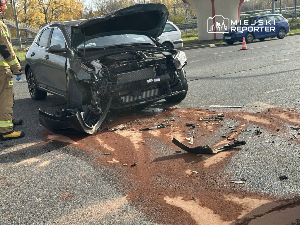 Wypadek drogowy z uszkodzonym samochodem osobowym i rozrzuconymi fragmentami na jezdni, obok strażak w mundurze.