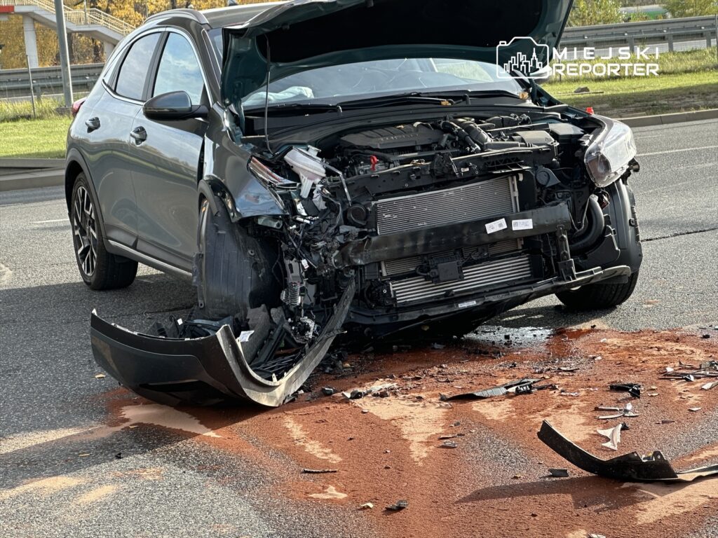 Uszkodzony samochód osobowy z otwartą maską leży na drodze, obok widać plamę płynu i fragmenty zderzaka.