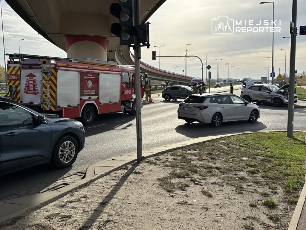Na skrzyżowaniu zajezdżają pojazdy ratunkowe, a przy uszkodzonych autach pracują strażacy.