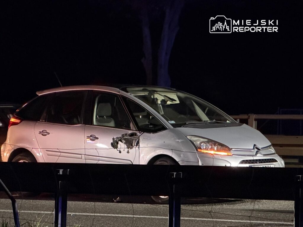 Uszkodzony biały samochód osobowy zaparkowany na poboczu drogi, z widocznym uszkodzeniem bocznej części.