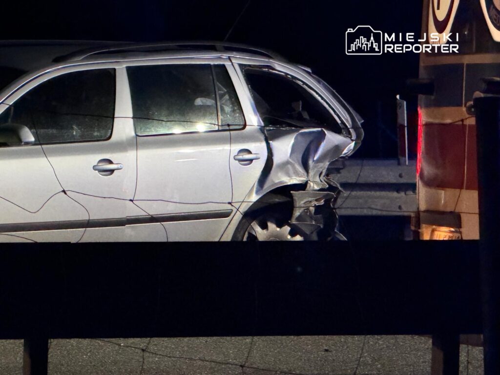 Uszkodzony samochód osobowy z wgniecioną tylną częścią stoi na poboczu drogi, obok niego widać pojazd służb ratunkowych.